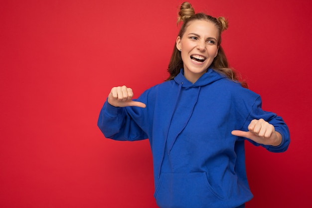 Jovem fascinante linda atraente positiva feliz alegre engraçada sorridente loira com dois chifres com emoções sinceras, vestindo um capuz azul moderno hippie isolado sobre um fundo vermelho com