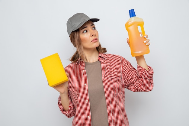 Jovem faxineira com camisa xadrez e boné segurando um frasco de material de limpeza e uma esponja, parecendo confusa, tendo dúvidas em pé sobre um fundo branco