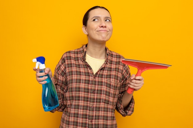 Jovem faxineira com roupas casuais segurando spray de limpeza e esfregão olhando para o lado sorrindo feliz e positiva