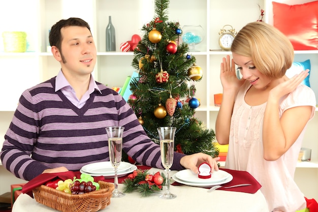 Jovem faz proposta de casamento com uma garota à mesa perto da árvore de Natal