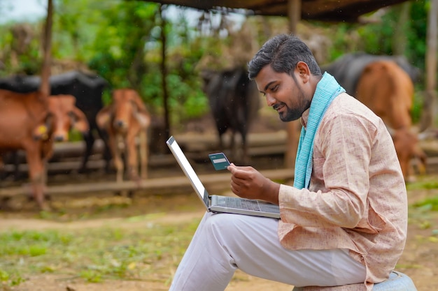 Jovem fazendeiro indiano usando laptop e cartão em sua fazenda
