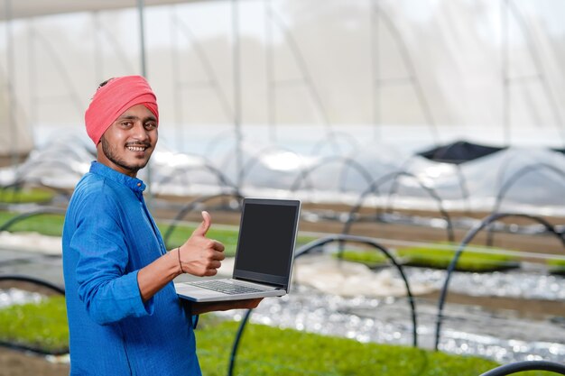 Jovem fazendeiro indiano usando laptop em uma estufa ou casa poli