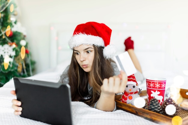 Jovem fazendo compras on-line no laptop fazendo pedidos no celular Garota deitada embaixo do Natal decorado