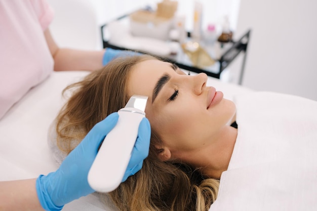 Jovem fazendo procedimento de beleza em esteticista de salão de spa usando dispositivo de peeling ultra-sônico clining