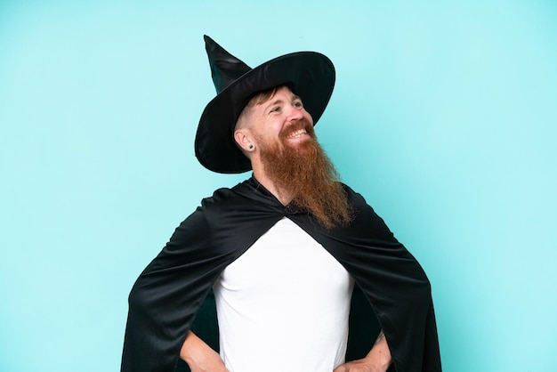 Jovem feiticeiro no halloween isolado em fundo azul posando com braços no quadril e sorrindo