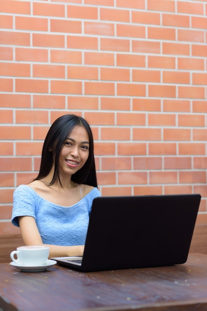 Jovem feliz adolescente asiática sorrindo com laptop e cappuccino na mesa de madeira contra a parede de tijolos