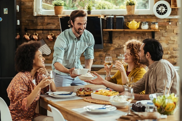 Jovem feliz almoçando com seus amigos e servindo comida na mesa de jantar