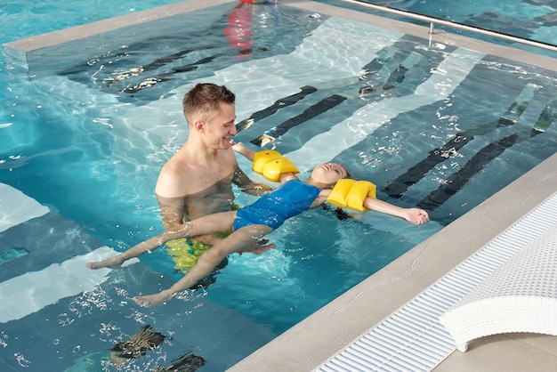 Jovem feliz apoiando sua filha com mangas de segurança durante a natação na piscina com água transparente em um moderno centro de spa