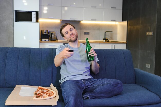 Foto jovem feliz bebendo cerveja e comendo pizza enquanto assiste a um jogo na televisão em casa