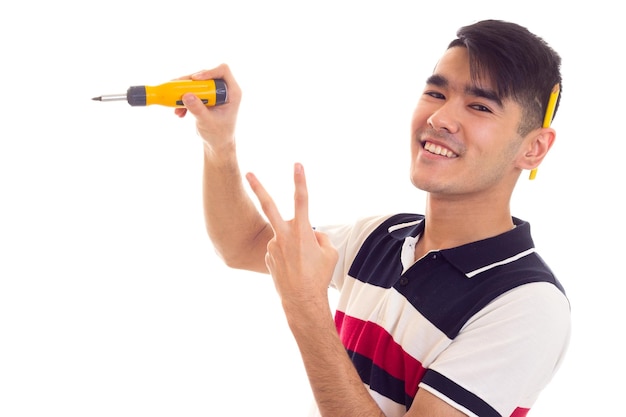 Jovem feliz com cabelo preto na camiseta branca e azul com lápis atrás da orelha segurando a chave de fenda amarela e mostrando a paz em fundo branco no estúdio.