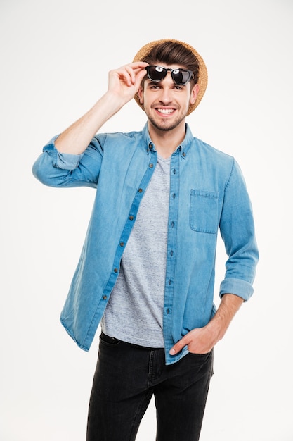 Jovem feliz com camisa azul e óculos escuros em pé e sorrindo sobre fundo branco