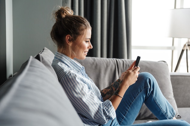 jovem feliz com camisa listrada sorrindo e usando o celular enquanto está sentada no sofá da sala de estar