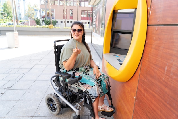 Jovem feliz com deficiência em cadeira de rodas usando caixa eletrônico e sorrindo para a câmera em um dia ensolarado de verão