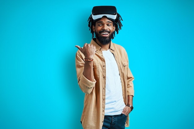 Jovem feliz com óculos de realidade virtual apontando para o espaço da cópia e sorrindo em pé contra a parede azul