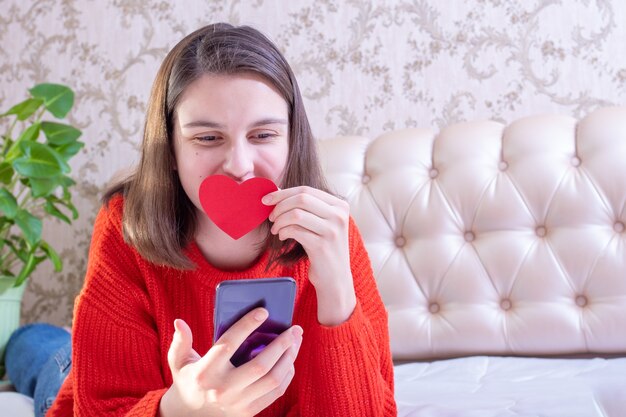 Jovem feliz com uma camisola vermelha, segurando um coração de papel vermelho e telefone, parabenizando família ou amigos no dia dos namorados em casa.