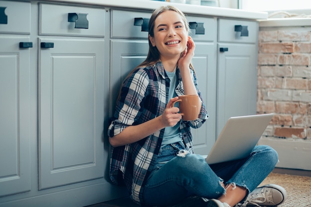 Jovem feliz com uma xícara na mão, sorrindo enquanto está sentada no chão com o gadget