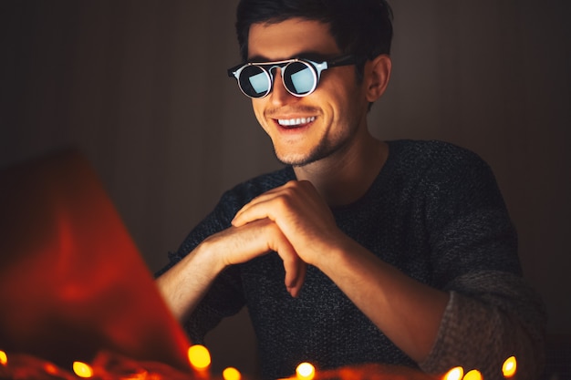 Jovem feliz de óculos redondos, olhando no laptop no quarto escuro com guirlandas em casa.