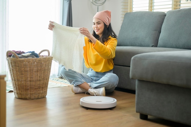 Jovem feliz dobrando roupas na sala de estar enquanto aspirador de pó robótico trabalhando