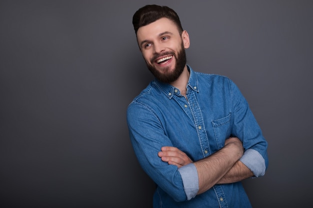 Jovem feliz e animado barbudo em jeans com os braços cruzados posando sobre uma superfície cinza