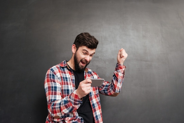 Jovem feliz e animado usando telefone celular e comemorando o sucesso em cinza