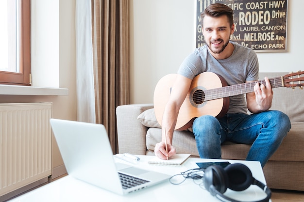 Jovem feliz e inspirado com um laptop tocando guitarra e escrevendo uma nova música no bloco de notas