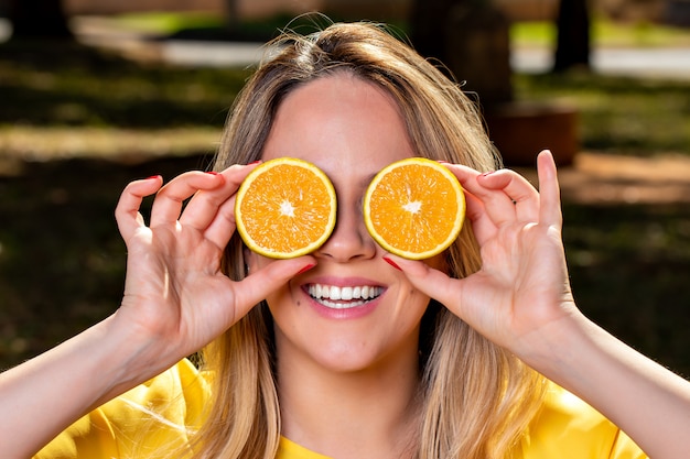 Jovem feliz e sorridente, brincando com uma laranja cortada ao meio e na frente de seus olhos. Foto no parque ao ar livre e sob árvores.