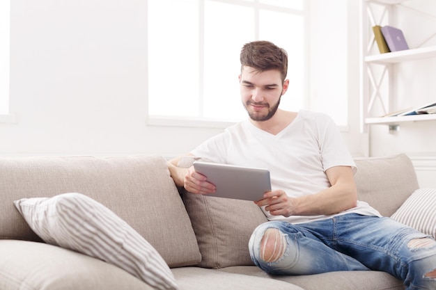 Jovem feliz em casa lendo em um tablet no sofá bege de uma sala iluminada, copie o espaço