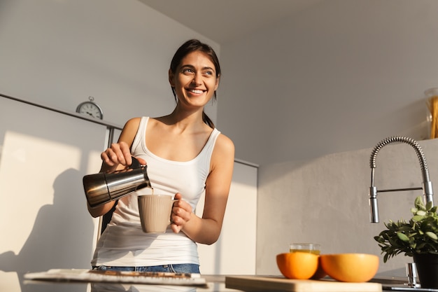 Foto jovem feliz em pé na cozinha pela manhã, tomando uma xícara de café