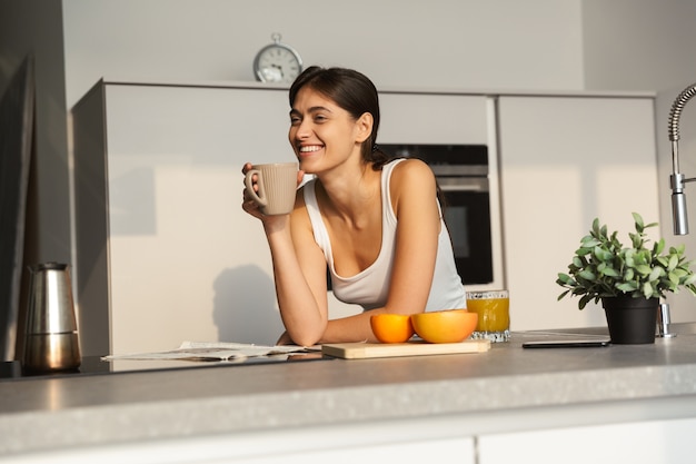 Jovem feliz em pé na cozinha pela manhã, tomando uma xícara de café