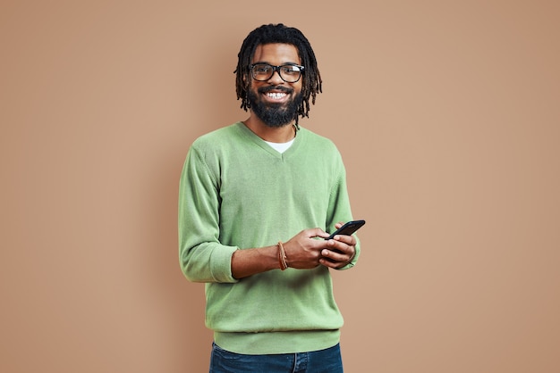 Jovem feliz em roupas casuais inteligentes usando um telefone inteligente em pé contra uma parede marrom