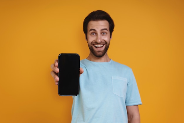 Jovem feliz em roupas casuais, mostrando seu telefone inteligente e sorrindo