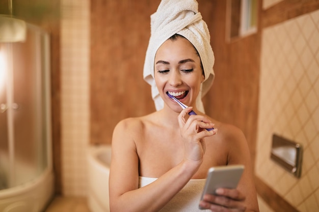 Jovem feliz escovando os dentes enquanto mensagens de texto no celular no banheiro