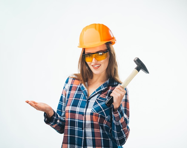 Jovem feliz garota atraente na construção de capacete laranja e óculos com martelo. Foto de estúdio totalmente isolada de artesã emocional.