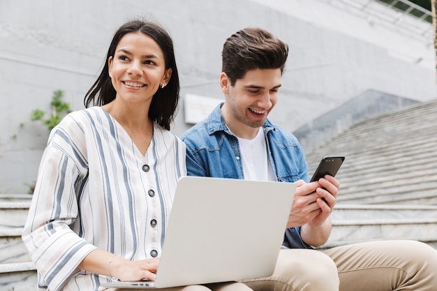 jovem feliz incrível casal amoroso empresários colegas ao ar livre do lado de fora nas etapas usando telefone celular e computador portátil.