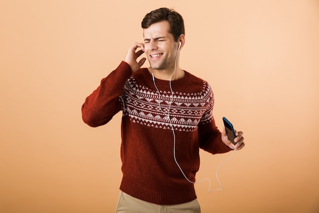 Jovem feliz isolado sobre uma parede bege usando telefone, ouvindo música por fones de ouvido e cantando