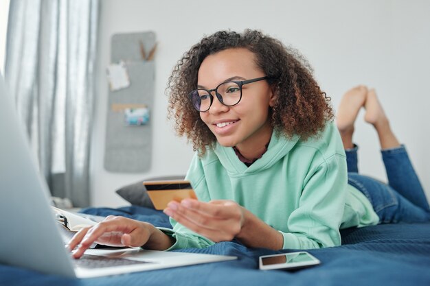 Foto jovem feliz mestiça de óculos e roupas casuais, deitada na cama em frente ao laptop e folheando as mercadorias na loja online