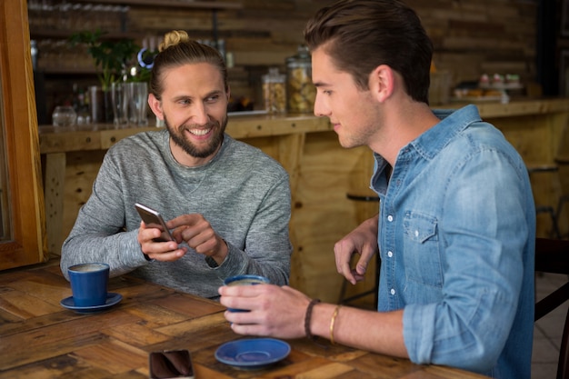 Jovem feliz mostrando telefone inteligente para um amigo na cafeteria