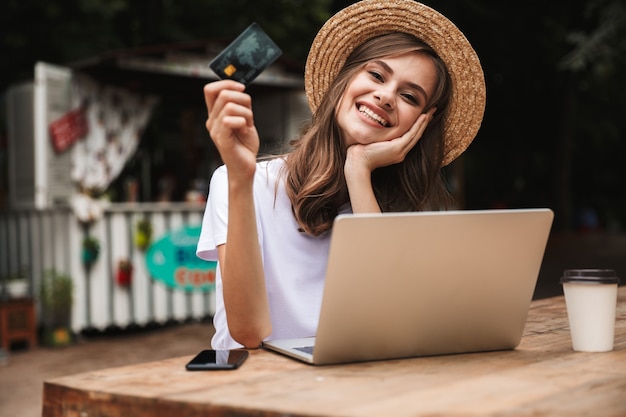 Jovem feliz mostrando um cartão de crédito de plástico enquanto está sentado com um laptop e tomando café no café ao ar livre