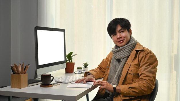 Jovem feliz na jaqueta amarela sentado no escritório em casa moderno e sorrindo para a câmera.