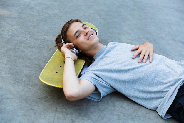 Jovem feliz passando um tempo na pista de skate, ouvindo música com fones de ouvido, deitado no skate