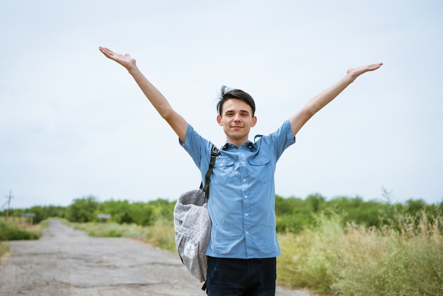 Jovem feliz posando com os braços erguidos em pé na estrada e olhando para a distância cara feliz com uma camisa azul e uma mochila de estudante livre aproveita as férias