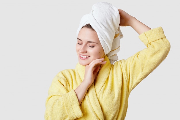 Jovem feliz relaxada tem pele macia, veste roupão amarelo e toalha na cabeça, toca a bochecha, tem tratamentos de beleza