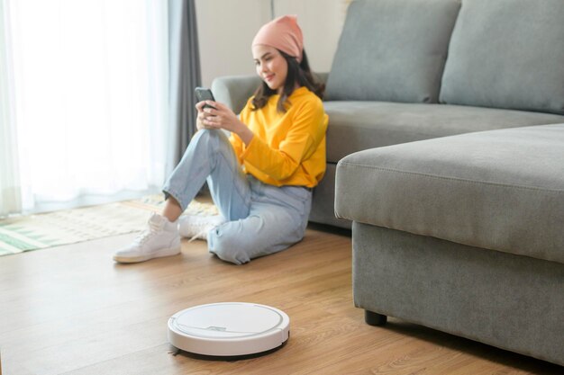 Jovem feliz relaxando e usando smartphone na sala de estar enquanto aspirador robótico trabalhando