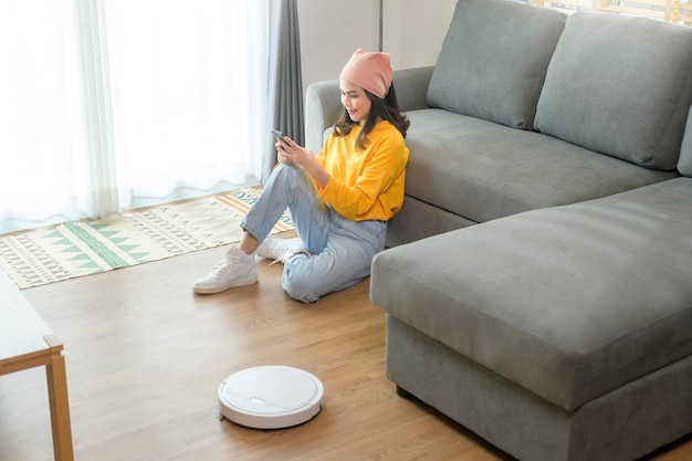 Jovem feliz relaxando e usando smartphone na sala de estar enquanto aspirador robótico trabalhando