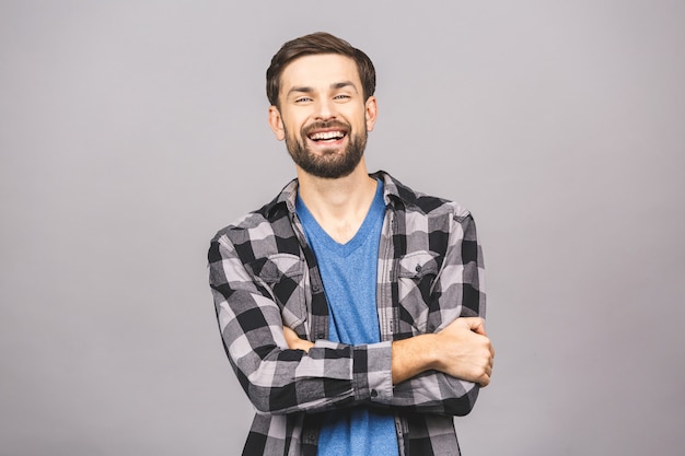 Jovem feliz. Retrato de jovem bonito na camisa casual, mantendo os braços cruzados e sorrindo em pé contra a parede cinza cinza.