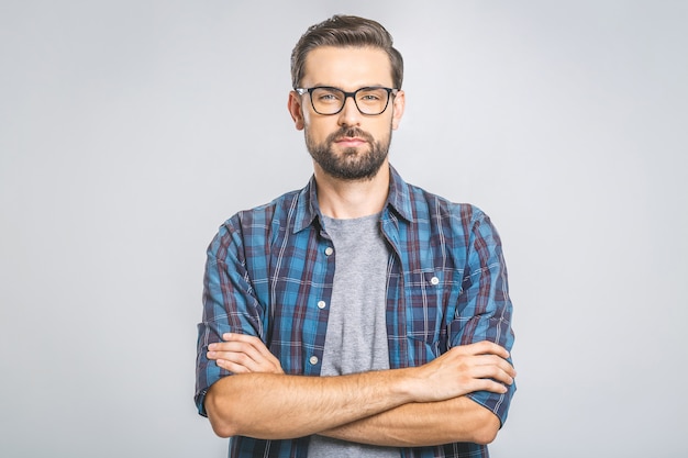 Jovem feliz. Retrato de jovem bonito na camisa casual, mantendo os braços cruzados e sorrindo em pé contra a parede cinza