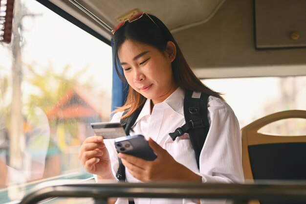 Jovem feliz segurando cartão de crédito e usando telefone celular enquanto está sentado dentro do transporte público de ônibus