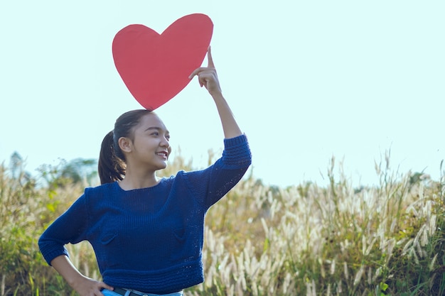 Foto jovem feliz segurando um coração de papel vermelho no prado. menina asiática