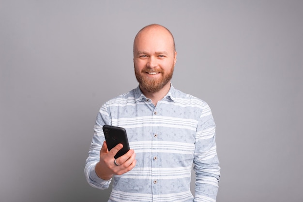 Jovem feliz segurando um smartphone, olhando para a câmera e sorrindo sobre um fundo cinza