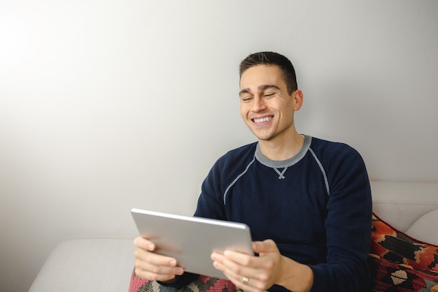 Jovem feliz segurando um tablet digital, sorrindo durante uma videochamada, sentado no sofá em casa